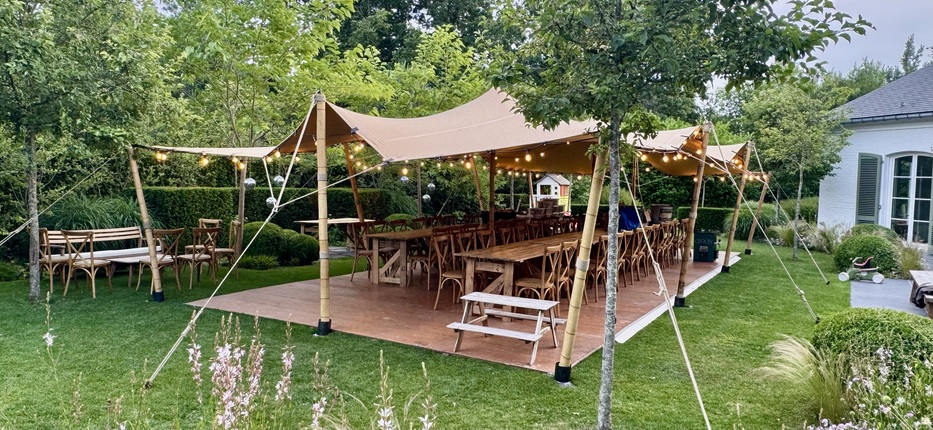 Tente nomade beige aménagée pour une réception dans un jardin, avec tables en bois et guirlandes lumineuses suspendues.